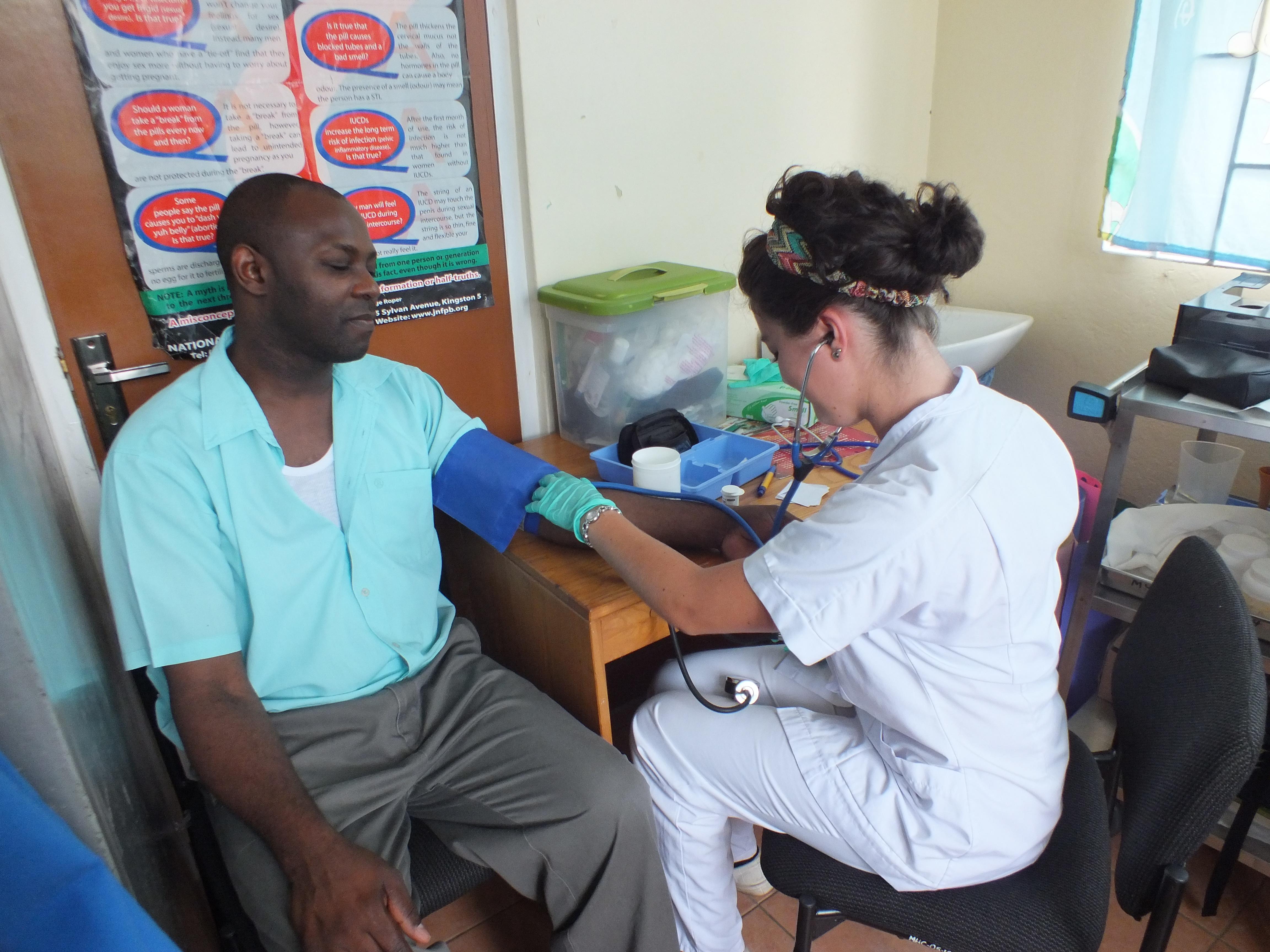 Un paciente realizándose un chequeo básico con una de nuestras internas de medicina en Jamaica. Interna de Medicina en Jamaica revisando la presión de un paciente.